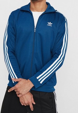 Hombre vistiendo una chaqueta Adidas azul con cremallera y rayas blancas en las mangas, y pantalones Adidas negros con rayas blancas, con las manos cruzadas al frente.