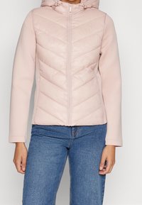 Ljusrosa puffjacka med huva, med chevronsöm, dragkedjestängning och långa ärmar, kombinerad med blå denimjeans.