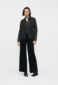 Jeune femme debout portant une veste en jean noire, un pantalon noir à jambes larges, un haut sombre et des bottines à talons noires brillantes.