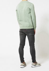 Felpa verde chiaro a girocollo, jeans slim-fit grigi scuri e sneakers beige con suole bianche. Cuciture e tasche visibili sui jeans.