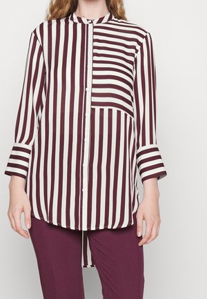 Femme portant une chemise à manches longues boutonnée avec des rayures verticales et horizontales bordeaux et blanches, associée à un pantalon uni bordeaux.