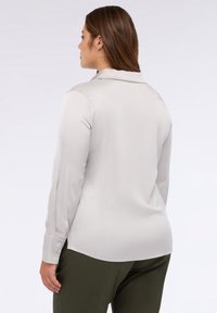 Blusa grigia chiara a maniche lunghe con una texture morbida, colletto, e dettagli di bottoni sulle maniche, abbinata a pantaloni verde oliva.