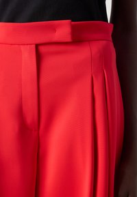 Rode geplooide broek met een zachte textuur, hoge tailleband en subtiele stiksels. Close-up weergave die de stof en het ontwerp benadrukt.