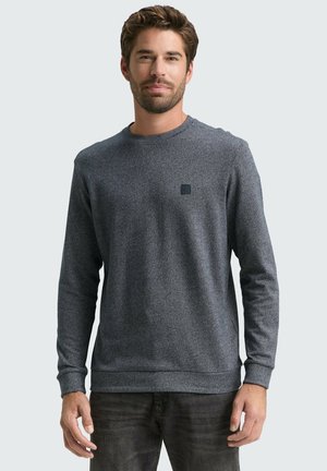 Grauer, strukturierten Sweatshirt mit Rundhalsausschnitt und langen Ärmeln. Mit einem kleinen schwarzen Logo auf der Brust. Kombiniert mit dunklen Jeans.