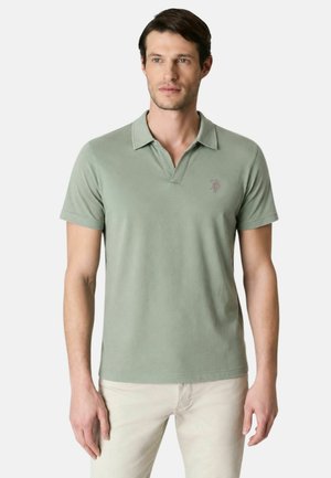 Uomo che indossa una polo verde chiaro a maniche corte e pantaloni beige chiaro, in piedi davanti a uno sfondo bianco uniforme.