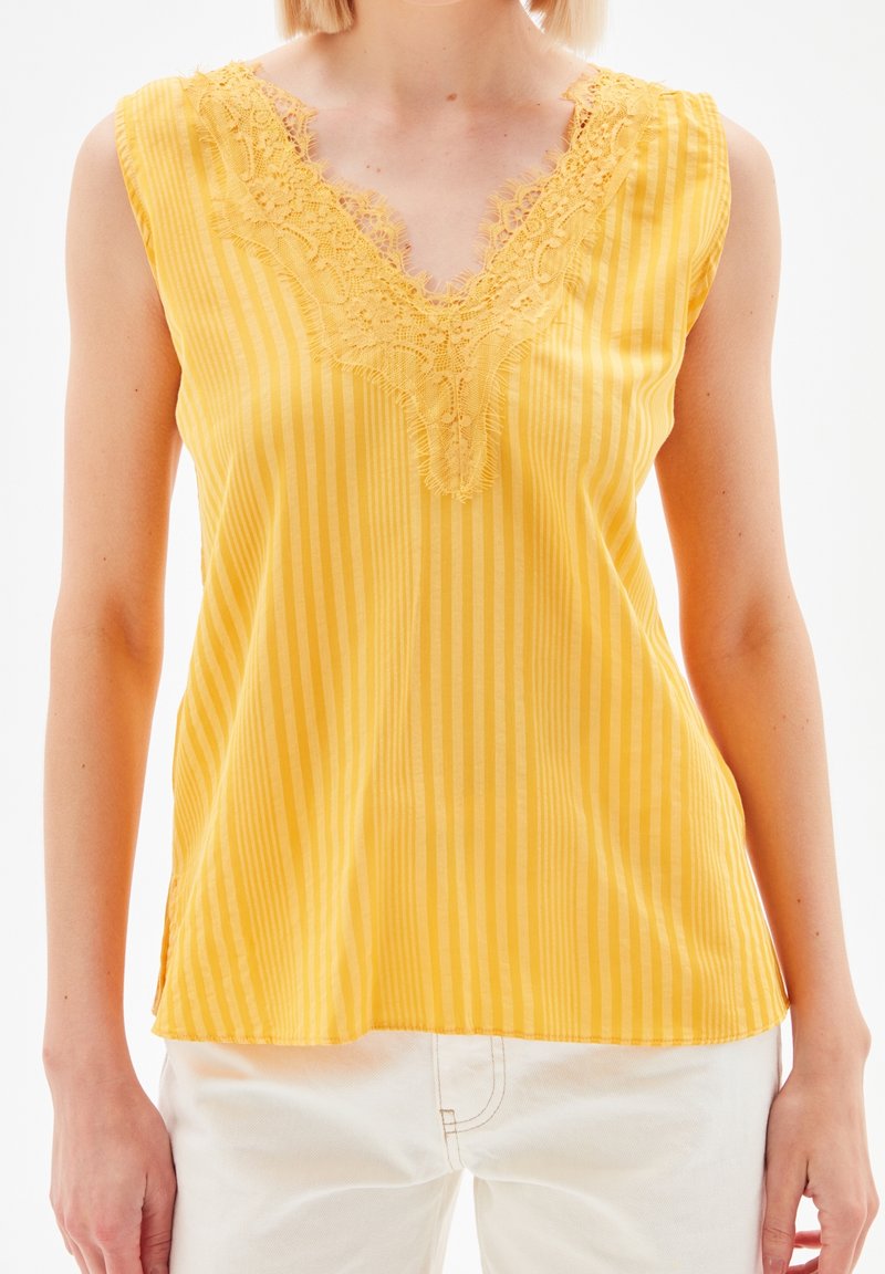 Blouse jaune sans manches à rayures verticales et col en V orné de dentelle portée avec un pantalon blanc sur une personne debout.