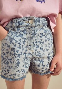 Shorts en denim ornés d'un motif de papillon bleu sur un fond clair, avec un ourlet effiloché et des poches avant. Fermeture à bouton en métal argenté.