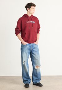 Maroon hoodie with white text, featuring a front pocket and drawstring hood. Pair of light blue ripped jeans and black sneakers.