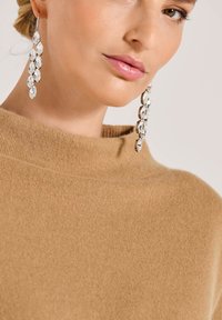 Nahaufnahme einer Frau, die hängende Kristallohrringe und einen camel-farbenen Strickpullover mit hohem Kragen trägt.