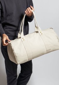 Beige canvas duffeltas met dubbele stoffen handvatten, ritssluiting en twee gouden hardwareaccenten aan de zijkant.