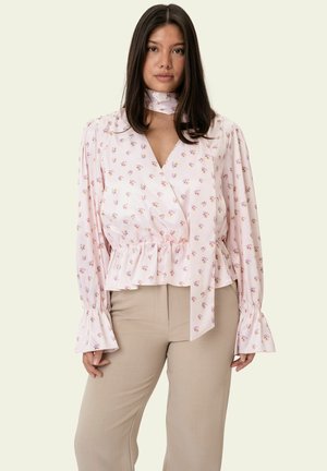 Naine, kes kannab heleroosat lillelist mähksepluusi pikkade varrukatega ja beeže pükse, seisab ühevärvilise heleda tausta ees.