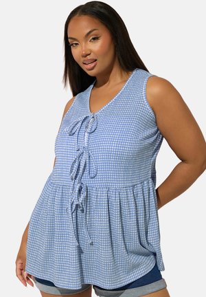 Frau trägt ärmelloses blau-weiß kariertes Oberteil mit drei vorderen Knoten und Jeansshorts, posiert mit einer Hand hinter dem Rücken.