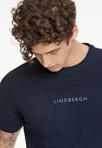 T-paita tummansinisestä puuvillasta, jossa on pyöreä kaula-aukko, lyhyet hihat ja vaaleansiniset "LINDBERGH" -logo rinnassa. Sileä rakenne.