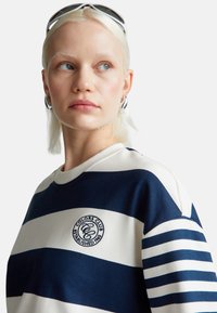 Felpa a righe navy e bianca con collo rotondo, caratterizzata da un logo ricamato circolare sul petto. Tessuto morbido e vestibilità comoda.
