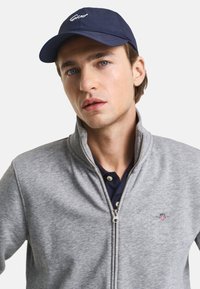Sudadera gris con cremallera y tejido texturizado, acompañada de una gorra de béisbol azul marino con logo bordado blanco. Diseño simple y casual.