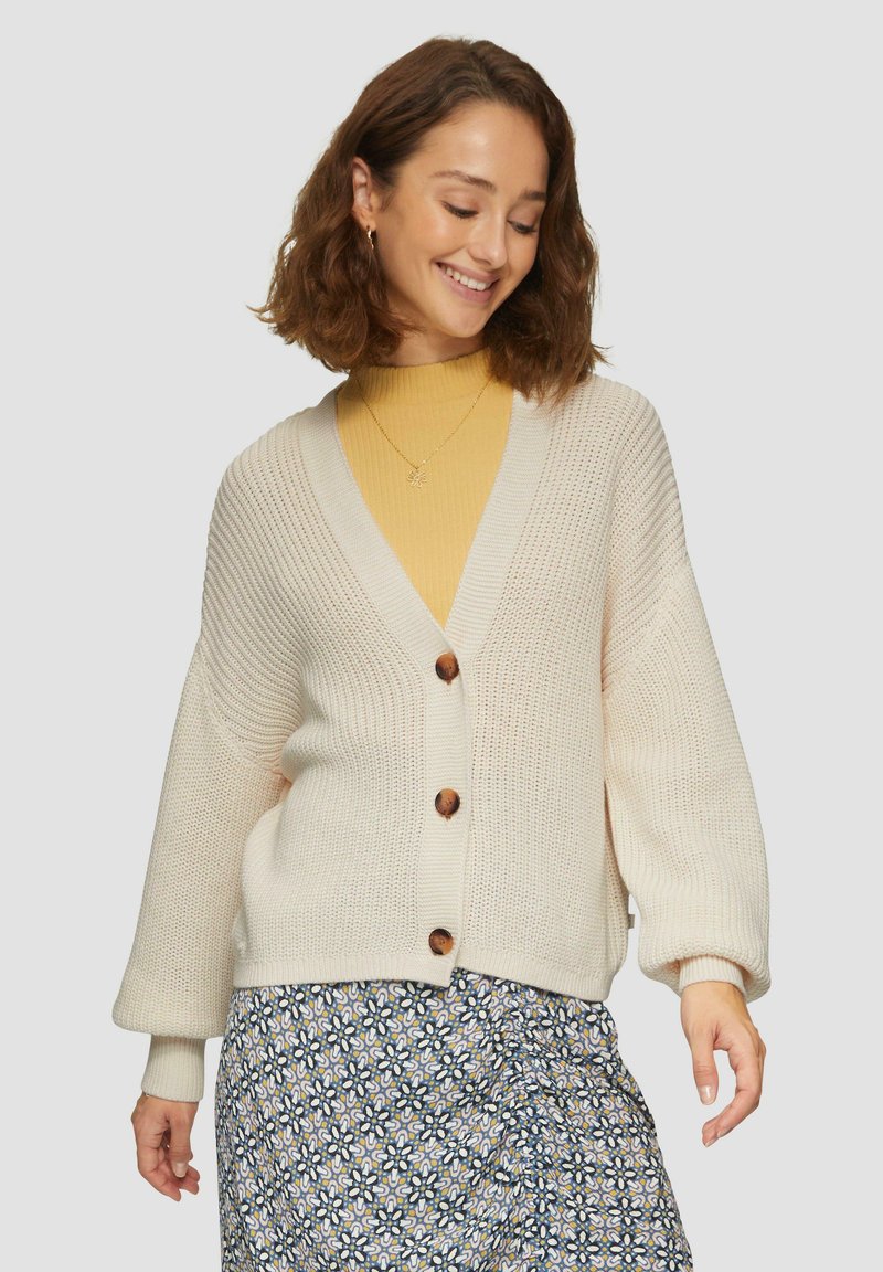 Cremefarbener Strickcardigan mit tiefem V-Ausschnitt, drei großen Holzknöpfen und weiten Ärmeln, getragen über einem gelben Rippentop.