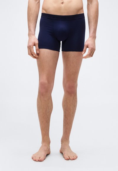 Άνδρας μοντέλο που φοράει μπλε μποξεράκια με λο γότυπο στη ζώνη, στέκεται ξυπόλητος μπροστά σε ένα απλό φωτεινό φόντο.