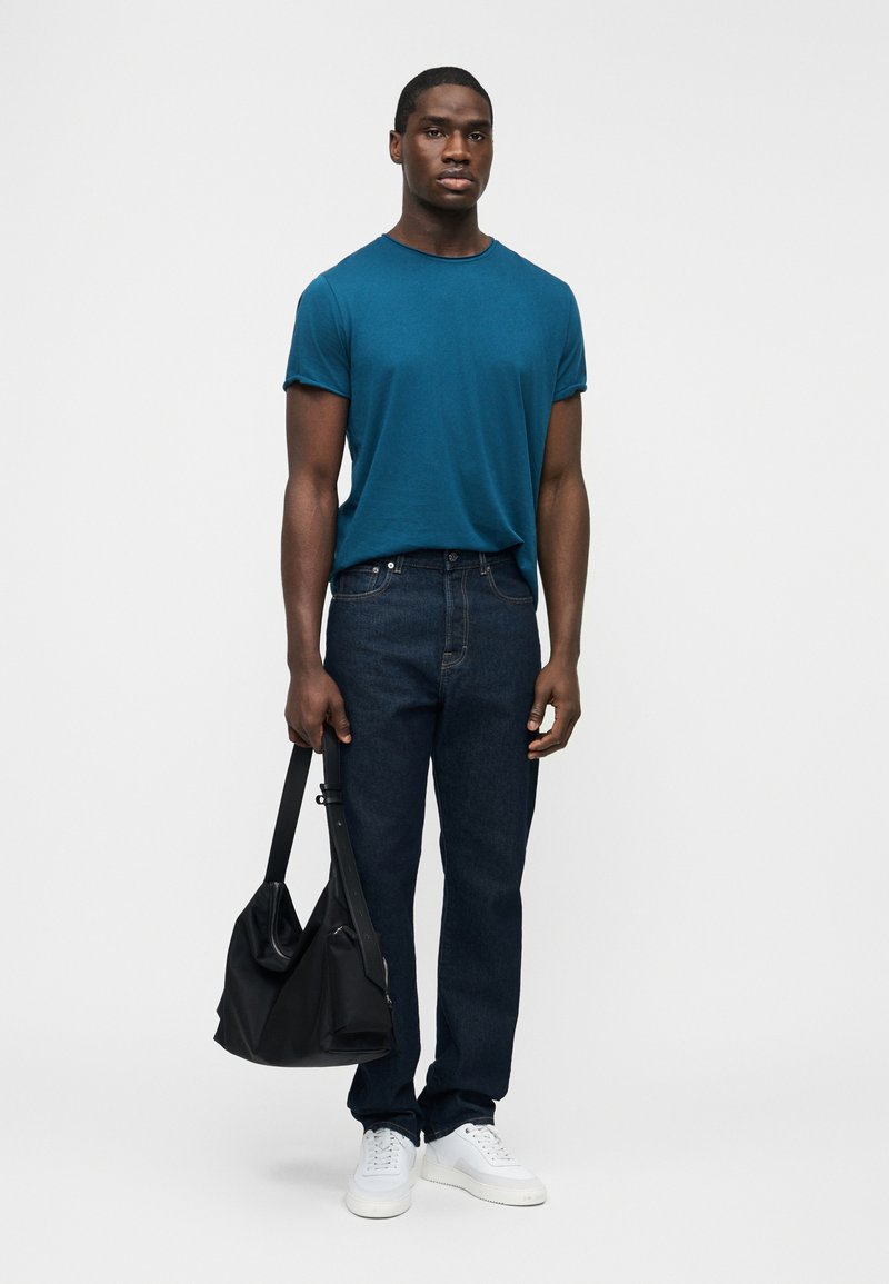 Man wearing teal t-shirt, dark jeans, and white sneakers, holding a black shoulder bag against a plain white background.
