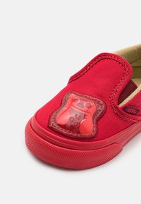 Zapato tipo mocasín rojo con material de lona, suela de goma y un motivo de oso de goma transparente en el empeine. Superficie texturizada y detalles de costura.