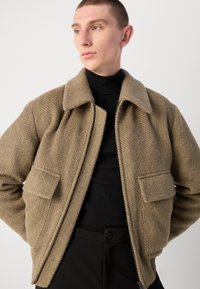 Hellbraune, strukturierte Jacke mit Fischgrätmuster, zwei Fronttaschen und Kragen, ergänzt durch einen schwarzen Rollkragenpullover darunter.