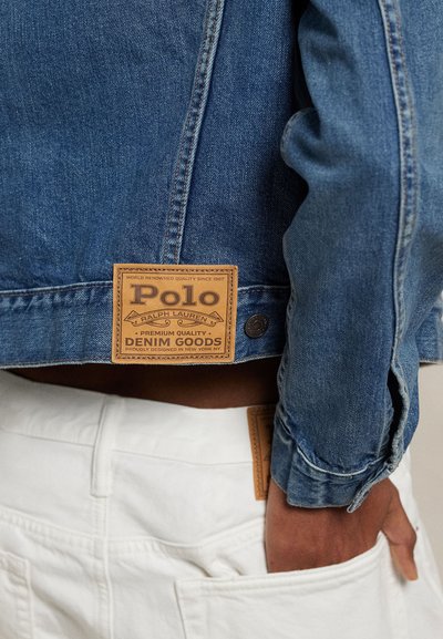 Primo piano di una persona che indossa una giacca di jeans blu Polo Ralph Lauren e pantaloni bianchi, con la mano appoggiata nella tasca posteriore.