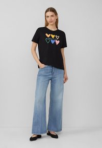 T-shirt noir en coton avec des motifs de cœurs multicolores, assorti à un jean large bleu clair et des chaussures plates noires.