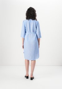 Light blue linen dress with three-quarter sleeves, belted waist, and a curved hem. Smooth texture, above-knee length. Brown block-heeled shoes.