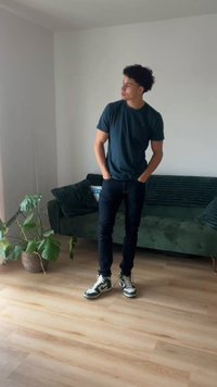 Jonge man in een zwart t-shirt en spijkerbroek staat in een woonkamer met een groene bank, kussens, houten vloer en een pot met een bladplant vlakbij.