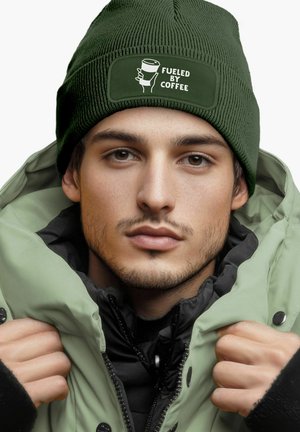 Green knit beanie with a rectangular patch that reads "FUELED BY COFFEE," worn with a light green insulated jacket.
