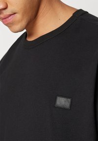 Close-up of a person wearing a black crew neck shirt with a small embroidered rectangular logo on the chest.