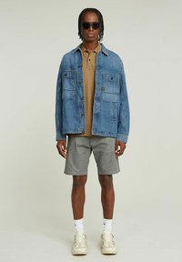 Giacca di jeans con due tasche sul petto, polo beige, pantaloni shorts grigi e sneakers di colore chiaro. Il modello indossa occhiali da sole neri.