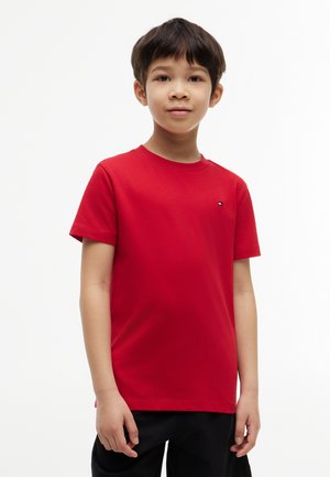 Ung dreng stående iført en ensfarvet rød kortærmet t-shirt og sorte bukser med et neutralt udtryk på en hvid baggrund.
