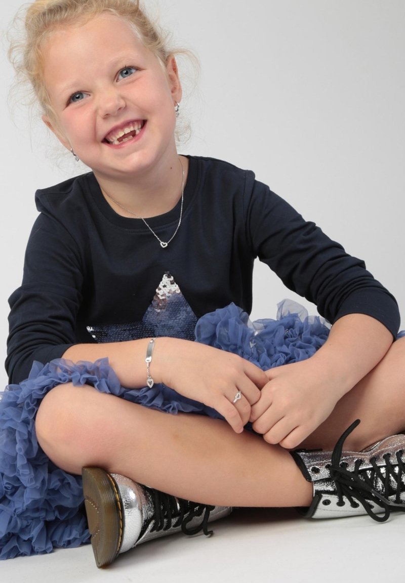 Kind in een marineblauwe lange mouw shirt met een zilveren ster, gelaagde blauwe gerimpelde rok, zilveren enkellaarsjes en zilveren sieraden.