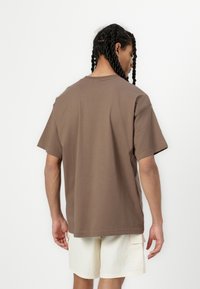 Brūns kokvilnas t-krekls ar brīvu piegriezumu, īsām piedurknēm, apaļo kakla izgriezumu un sānu šuvju detaļu, kombinēts ar gaišiem šortiem.