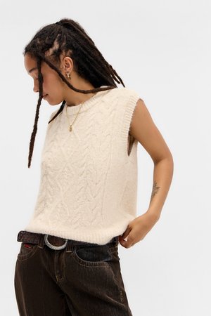 Knit sleeveless sweater in light beige with a textured cable pattern. Paired with dark denim jeans and a textured belt.