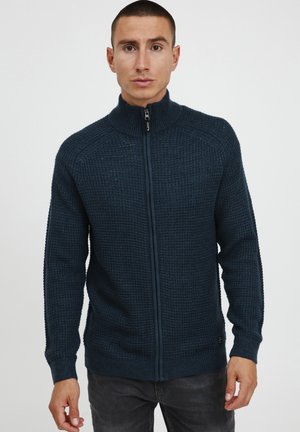 Sudadera de cremallera color azul marino con un patrón de punto texturizado, cuello alto y puños acanalados; presenta un pequeño logo en el lado inferior.