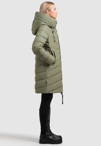 Olijf groen pufferjas met een hoge kraag, ritssluitingen en een rits aan de voorkant. Draag het met zwarte leggings en kniehoge zwarte laarzen.