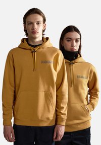 Dos modelos que llevan sudaderas amarillas de tejido suave. Cada una cuenta con un bolsillo delantero, detalles de logo y cordones con acentos metálicos.