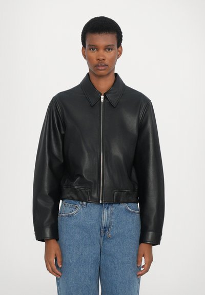 Chaqueta corta de cuero negro con cuello tipo solapa, cierre de cremallera y puños acanalados. Lleva encima unos vaqueros de mezclilla azul. Textura suave y diseño elegante.