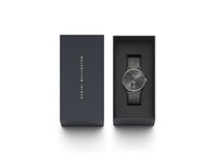 Reloj negro con correa de malla en una caja azul. El reloj presenta una esfera redonda, un dial oscuro y acentos plateados. La caja tiene letras doradas.