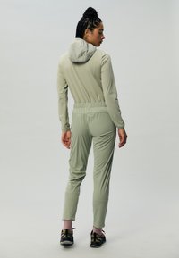 Jumpsuit lungo a maniche lunghe di colore verde chiaro, con cappuccio, vita elasticizzata e pantaloni affusolati. Il materiale appare liscio e traspirante. Sneakers nere.
