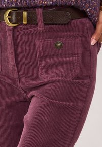 Corduroy broek in een rijke bordeauxkleur met verticale ribbels, voorzien van een subtiele zakdetail en een bruine leren riem met een messing gesp.