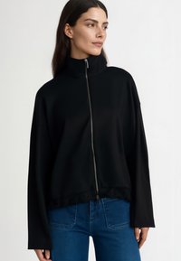 Črna jakna z zadrgo, visoki ovratnik, široki rokavi in teksturiran rob spodaj. Nosi se čez modre kavbojke, narejena iz gladkega, mehkega materiala.