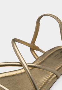 Χρυσό σανδάλι με λεπτά μεταλλικά λουριά που διασταυρώνονται πάνω από το πόδι και θηλιά γύρω από τη φτέρνα, σε λευκό φόντο.