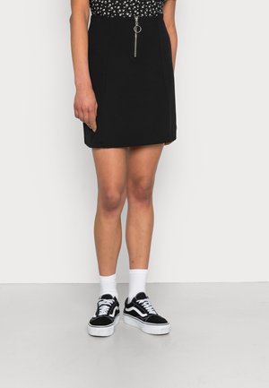 Saia mini preta com fecho éclair na frente e puxador de anel metálico, combinada com ténis em preto e branco. Textura suave e ajuste personalizado.