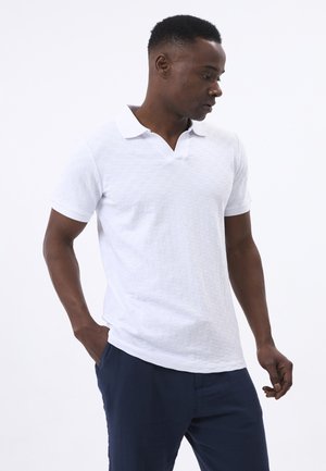 Camisa polo blanca con un diseño de patrón texturizado, con un collar clásico y mangas cortas, combinada con un pantalón azul oscuro.