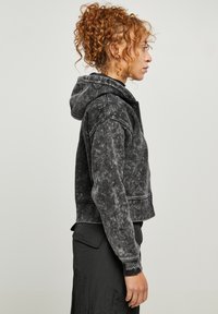 Sudadera negra recortada con un acabado texturizado, que cuenta con capucha, cierre de cremallera y costuras en contraste. La vista lateral resalta el ajuste.