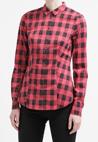 Camicia a quadri rossi e neri, con maniche lunghe, colletto e una forma aderente, caratterizzata da bottoni visibili e una tasca laterale.