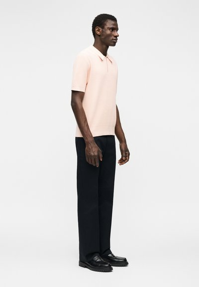 Άνδρας που φοράει ανοιχτό ροζ πόλο με κοντά μανίκια, μαύρο παντελόνι με ίσια μπατζάκια και μαύρα μοκασίνια, στέκεται με κατεύθυνση προς τα δεξιά σε απλό λευκό φόντο.