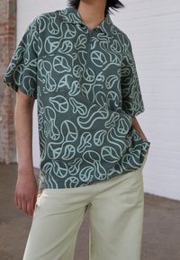 Camicia a maniche corte in tessuto verde scuro con motivi astratti di foglie verde chiaro, caratterizzata da un colletto e una tasca sul petto.